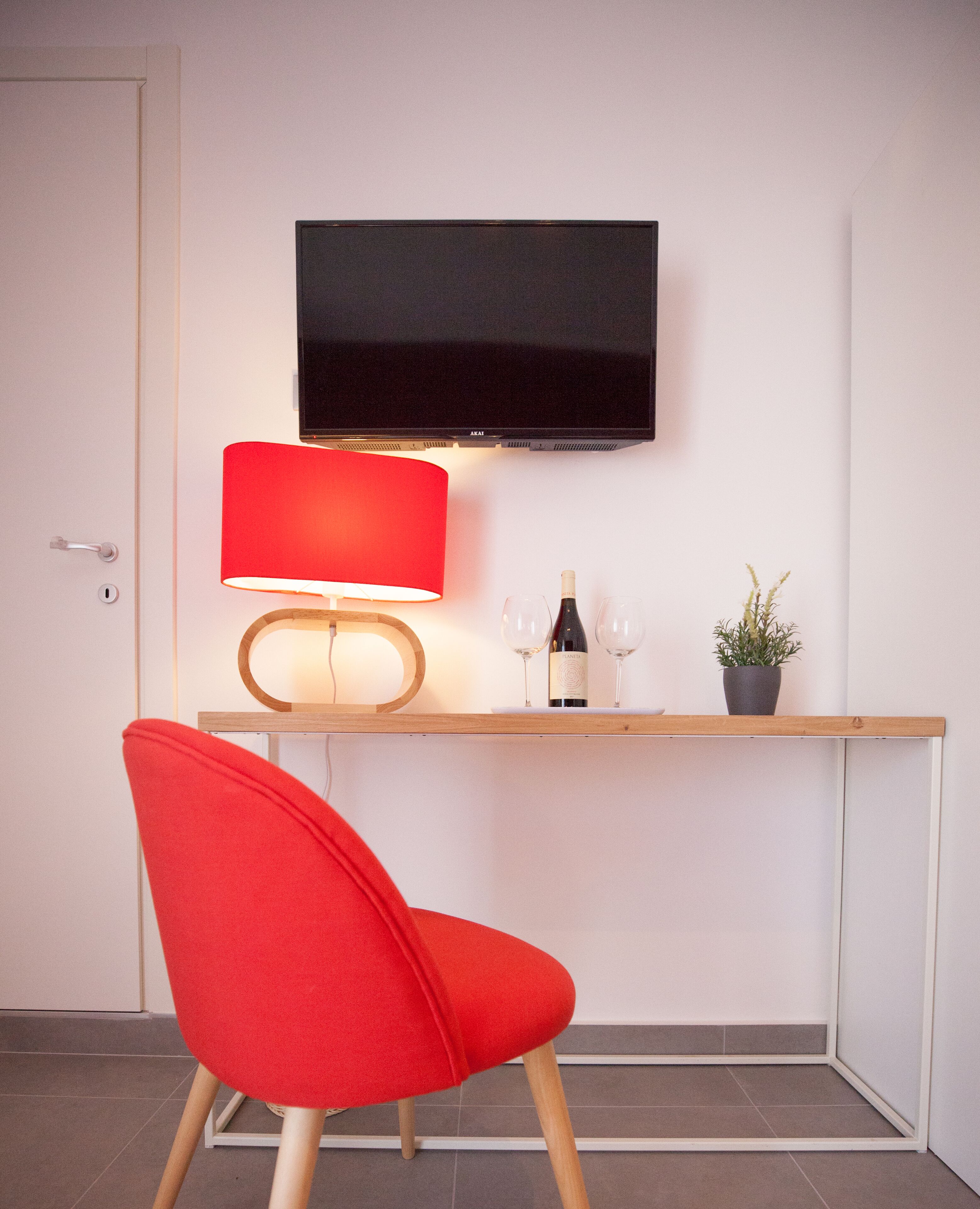 A modern room with a wooden desk, a red chair, a flat-screen TV, a red lamp, two wine glasses, a bottle of wine, and a potted plant.