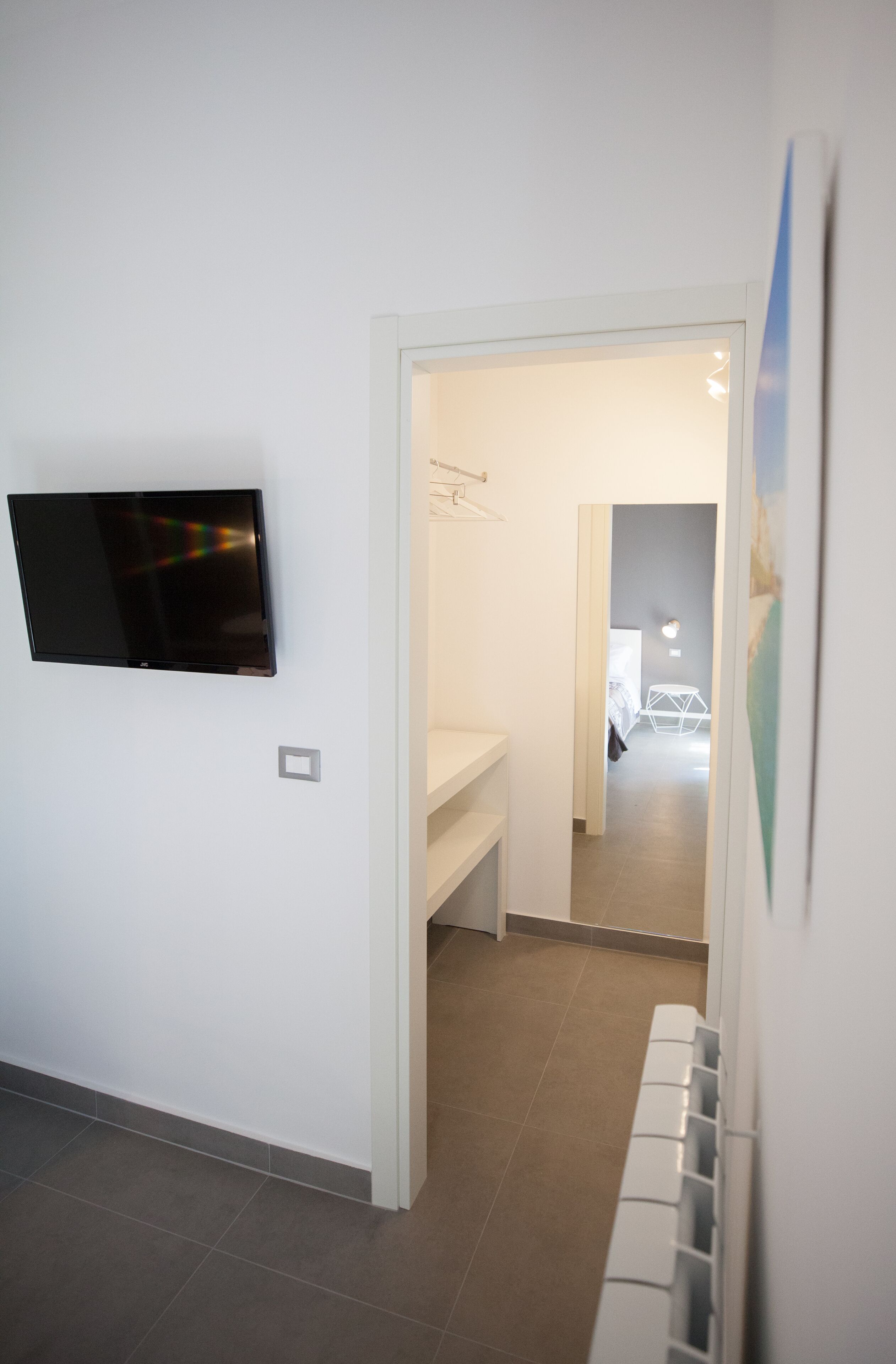 A hallway with a flat-screen TV mounted on the wall, a white bench, and a door leading to another room.