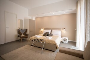 Un dormitorio moderno con una cama, mesitas de noche, una silla, un taburete pequeño y una ventana con cortinas.
