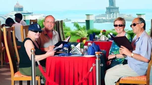 Un grupo de personas sentadas en una mesa con mantel rojo, vista a un paisaje urbano costero y una estatua al fondo.