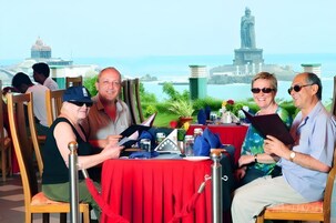 A group of people seated at a table with a red tablecloth, a view of a coastal cityscape, and a statue in the background.