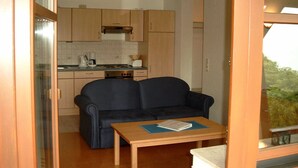 Una kitchenette compacta con estufa, cafetera y tostadora. Un sofá azul oscuro se encuentra frente a la kitchenette, con una pequeña mesa de madera a su lado.