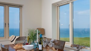 A dining table set with a bottle of wine, glasses, bread, and a candle, overlooking a scenic view of the ocean. - 5 The Vista, Newquay -  Walk to Beach - Free Parking & WiFi - Sea & Beach Views (Newquay)