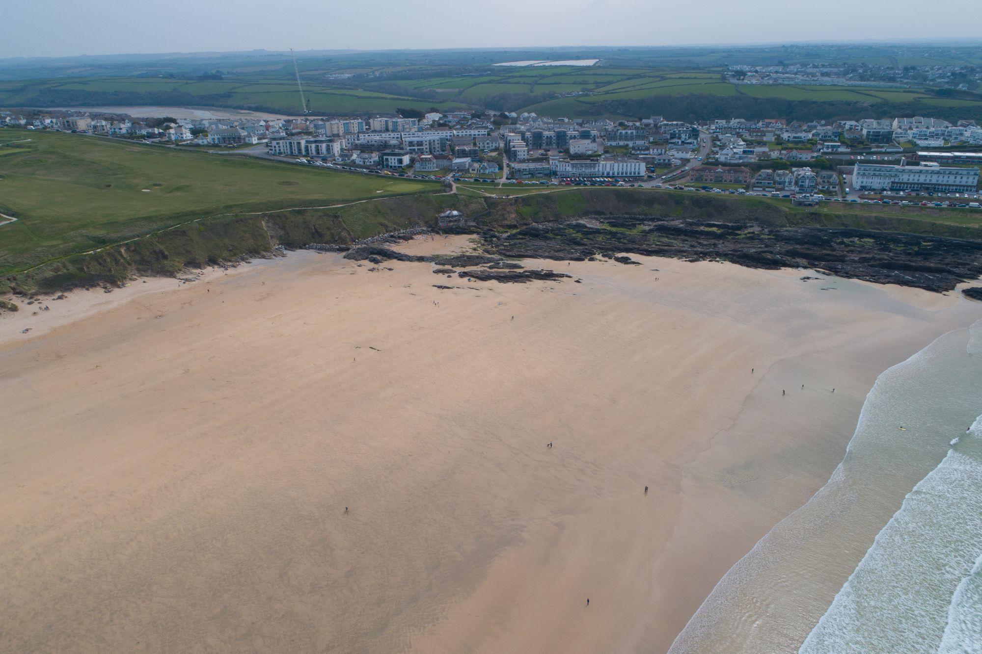 An aerial view of a coastal area with a sandy beach, a residential area with multi-story buildings, and a distant body of water.