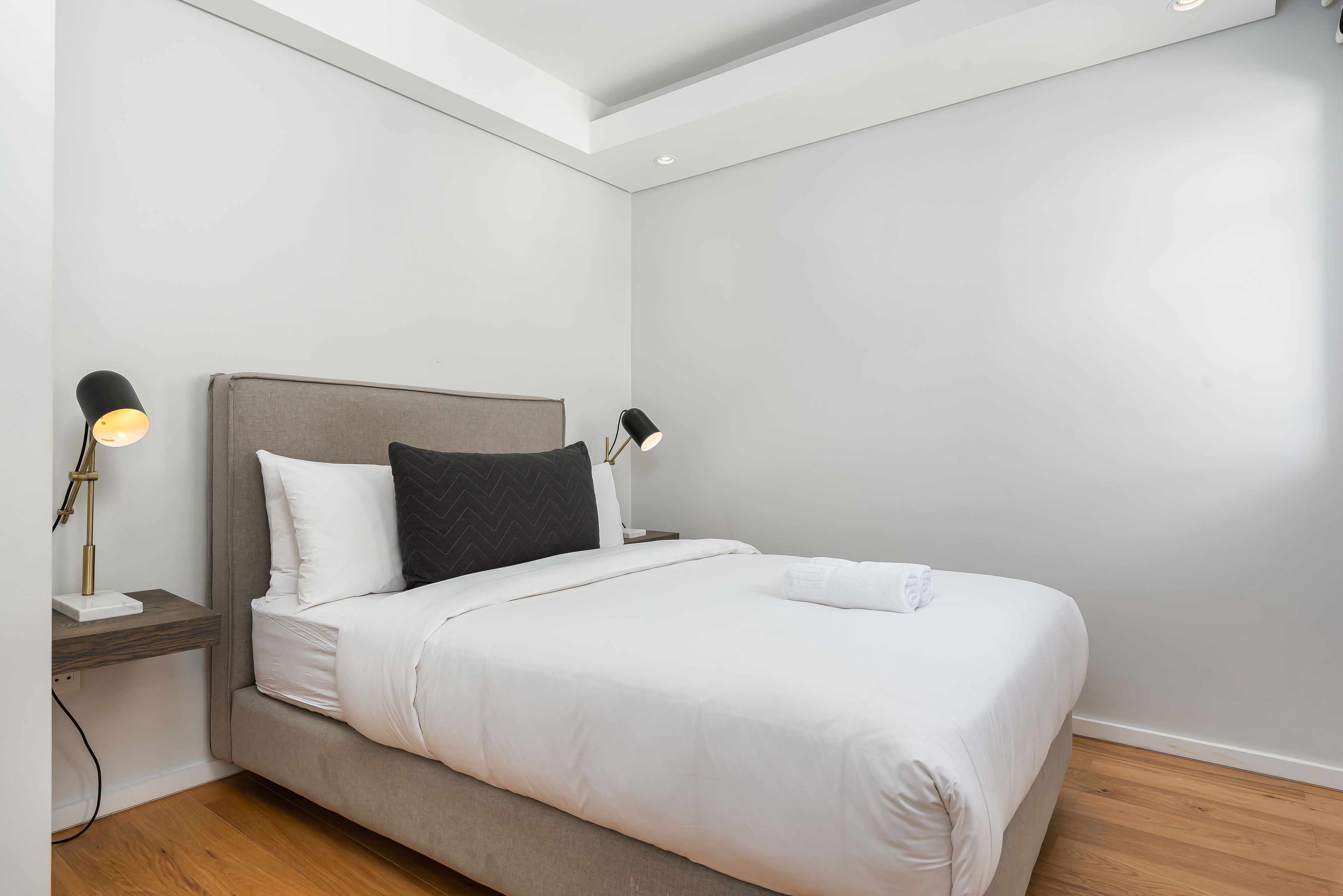 A neatly made bed with white linens, a grey headboard, and two bedside lamps.