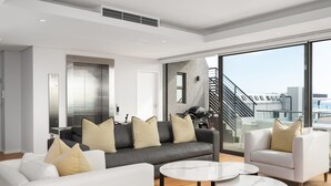 Un salon moderne avec un canapé gris, des fauteuils blancs et un mur en verre donnant sur la ville.