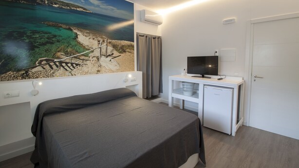 A bedroom with a bed, a desk with a television, a small refrigerator, and a large window with a coastal view. - Hostal Sa Vinya Suites (Sant Joan de Labritja)