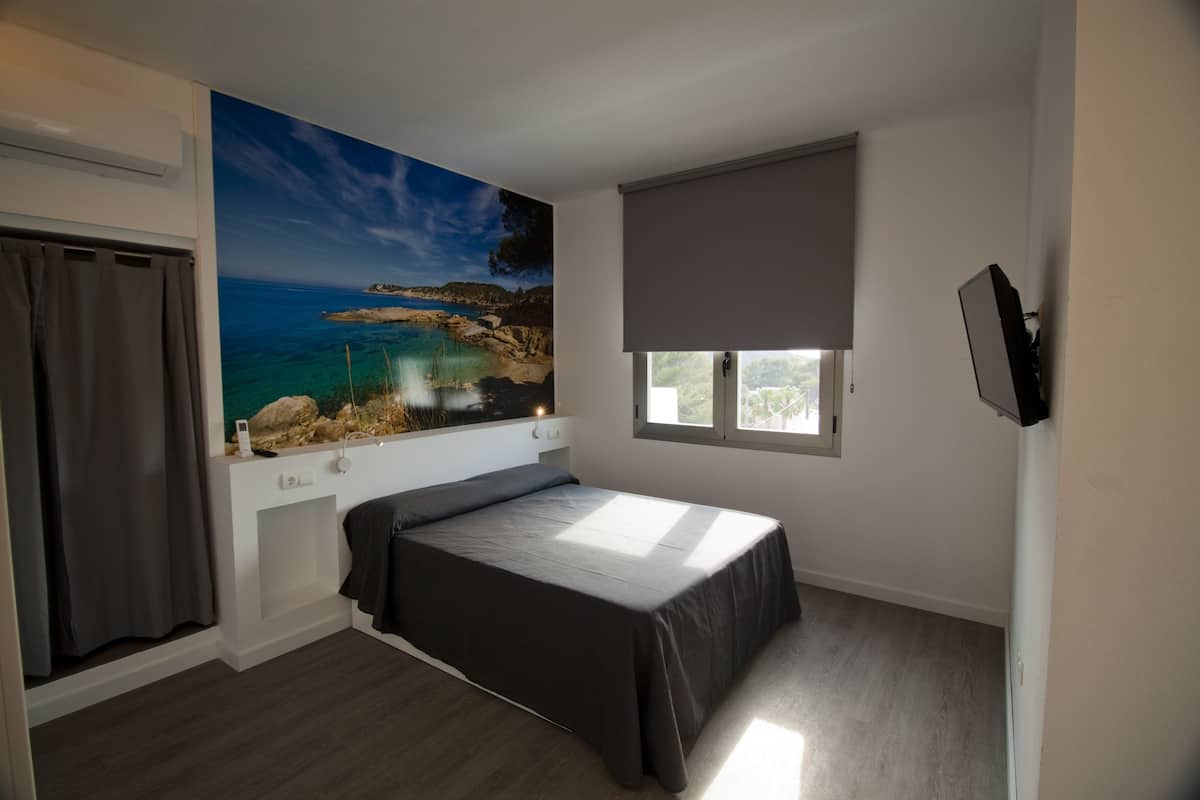 Un dormitorio con cama, ventana, un cuadro de una escena costera y televisor montado en la pared.