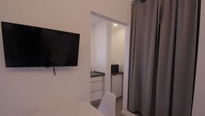 A modern room with a flat-screen TV mounted on the wall, a white chair, a small table, and a sliding door with curtains. - Hostal Sa Vinya Suites (Sant Joan de Labritja)