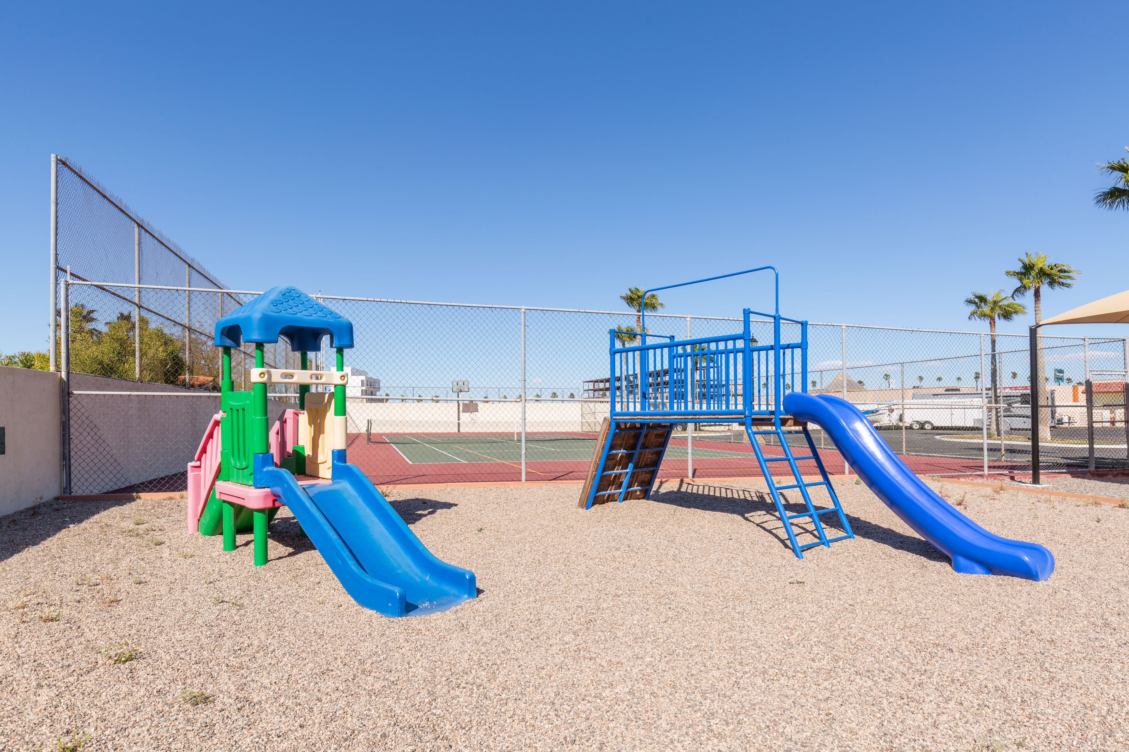 A playground with a blue slide and climbing structures, surrounded by a fence and palm trees.
