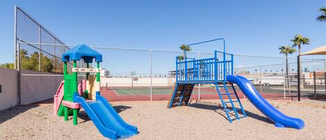 A playground with a blue slide and climbing structures, surrounded by a fence and palm trees.