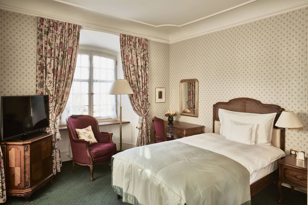 Ett hotellrum med en stor säng, en tv, en stol, ett skrivbord och ett fönster med gardiner.
