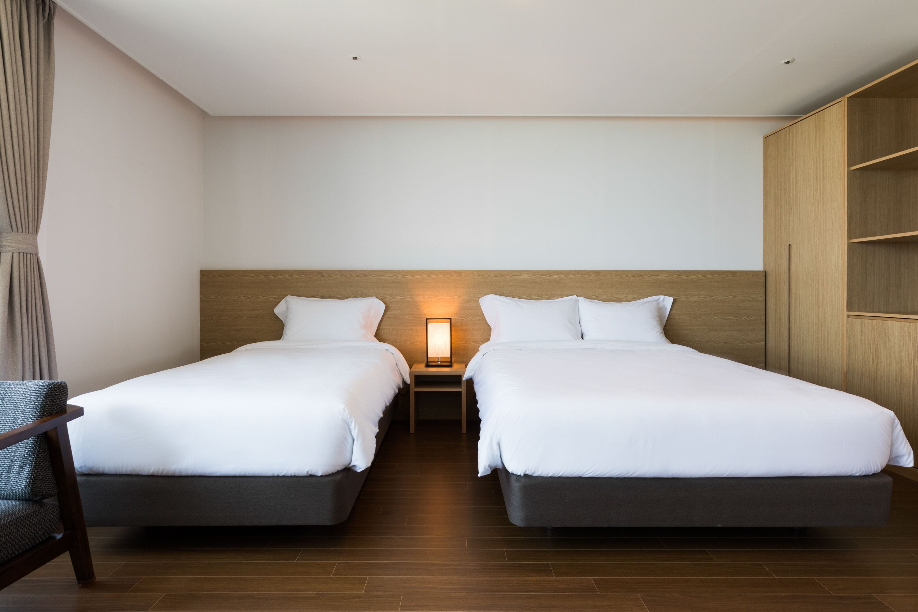 Habitación de hotel con dos camas, una mesita de noche con lámpara, un armario de madera y una silla.