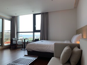 A hotel room with a large bed, a sofa, a chair, and a view of the city through large windows. - Lemain hotel (Jeju City)
