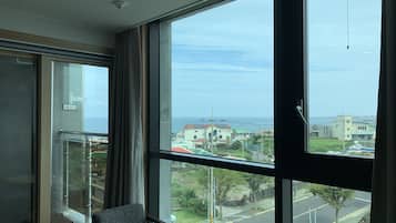 A room with a large window offering a view of a coastal area, a small table with a remote control, and a chair.
