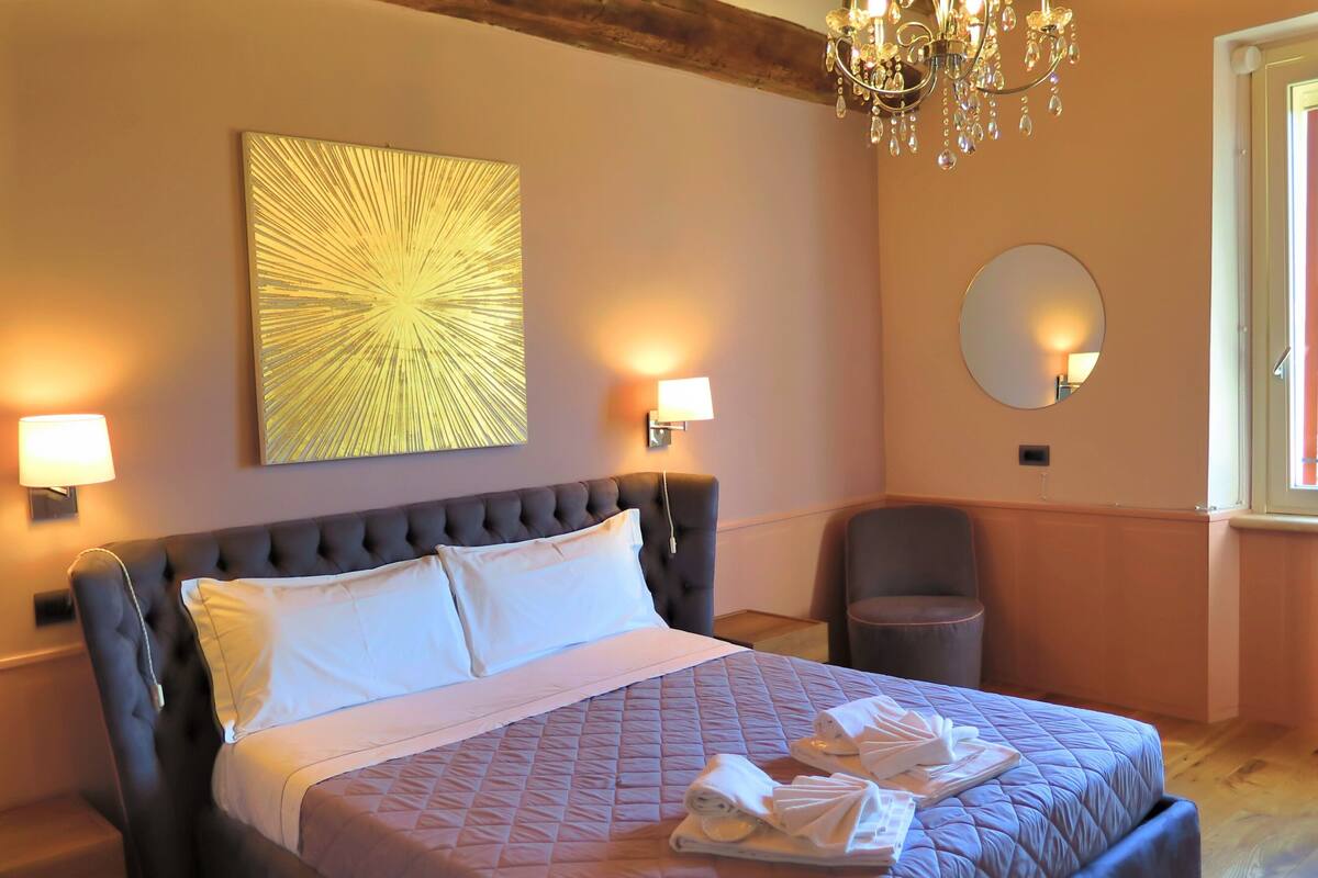 A hotel room with a large bed, two wall-mounted lamps, a chandelier, a round mirror, and a window with a view.