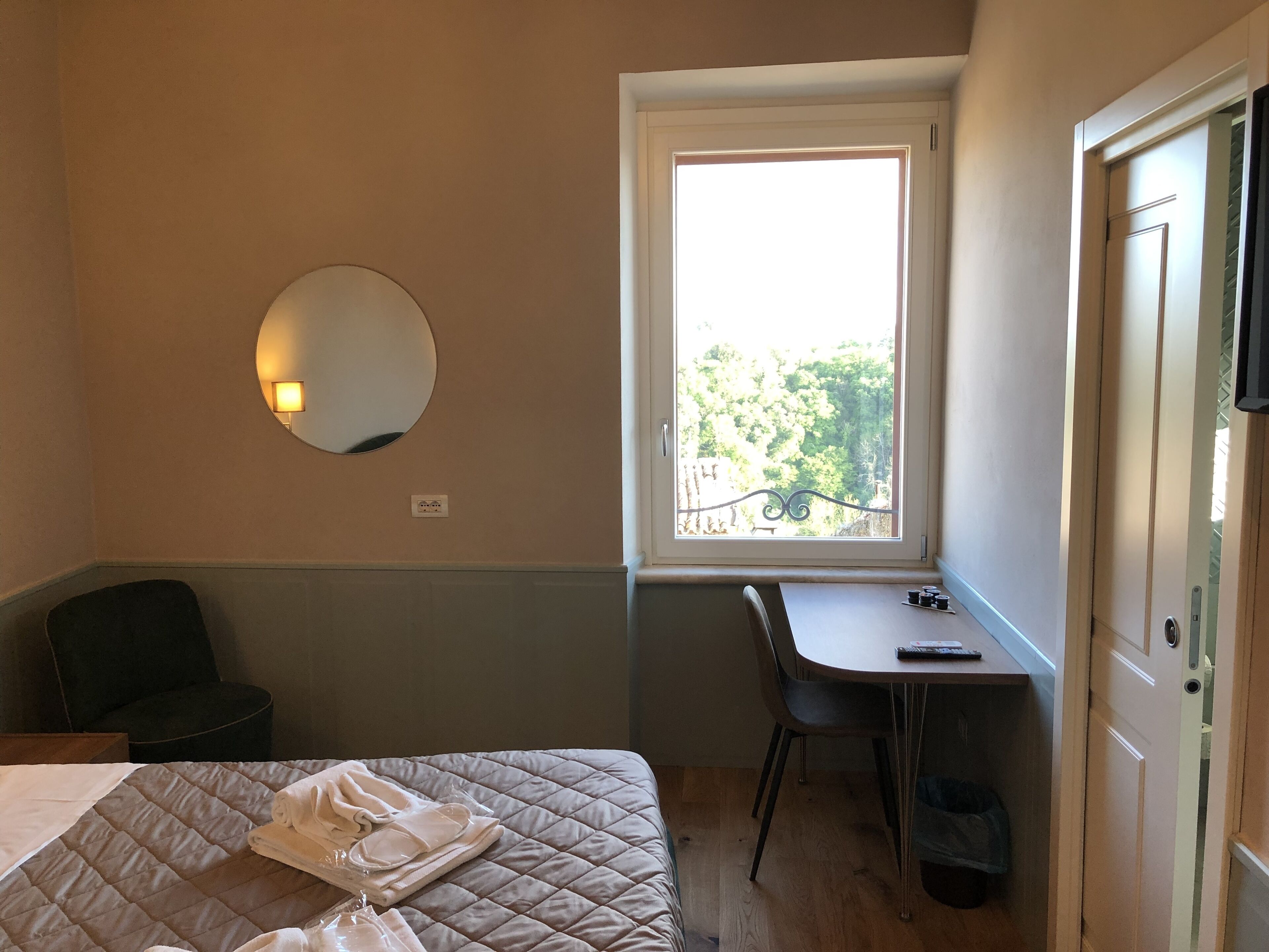 Een slaapkamer met een bed, een bureau, een stoel, een raam met uitzicht op bomen, en een deur.