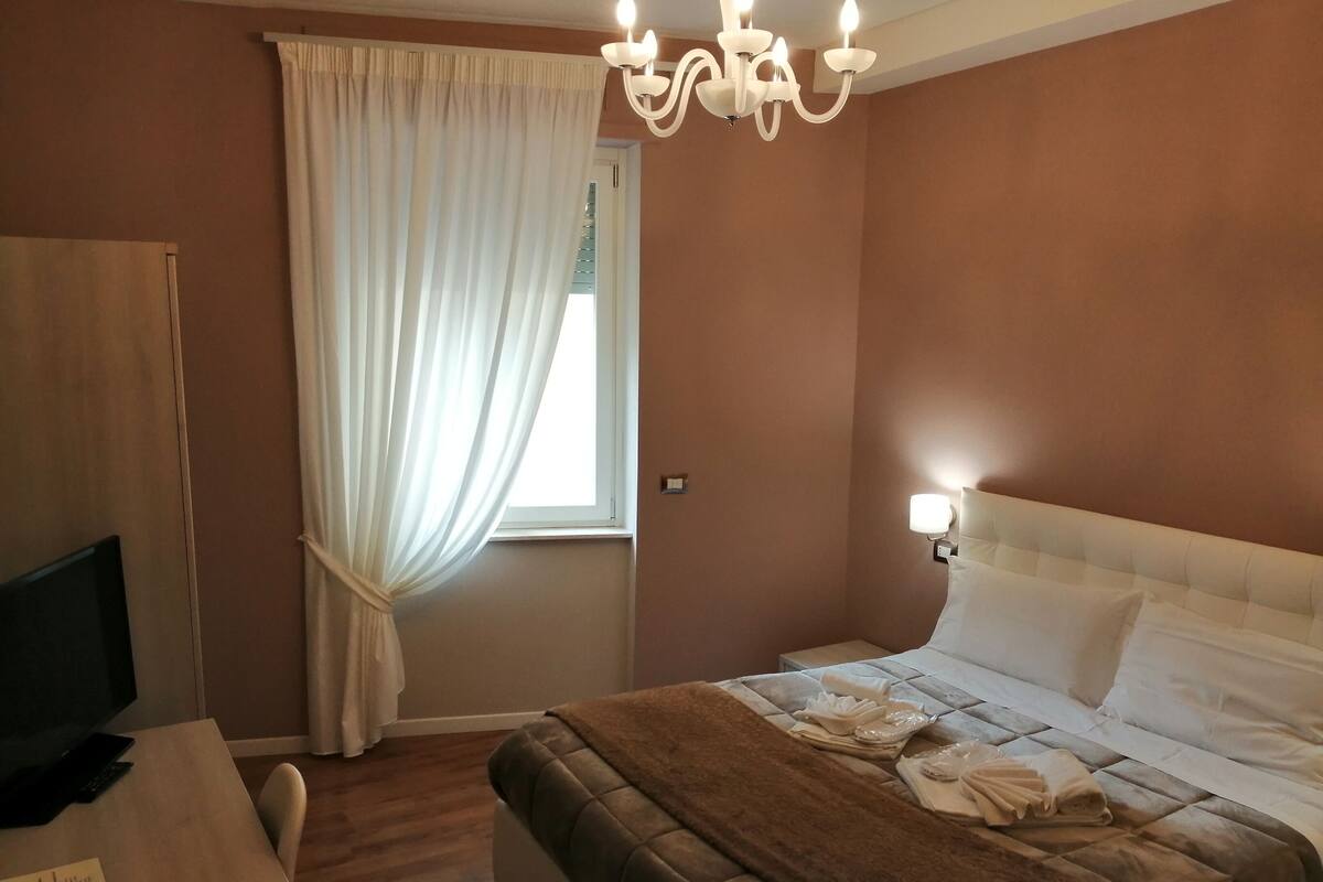 A hotel room with a bed, a TV, a desk, and a window with curtains.