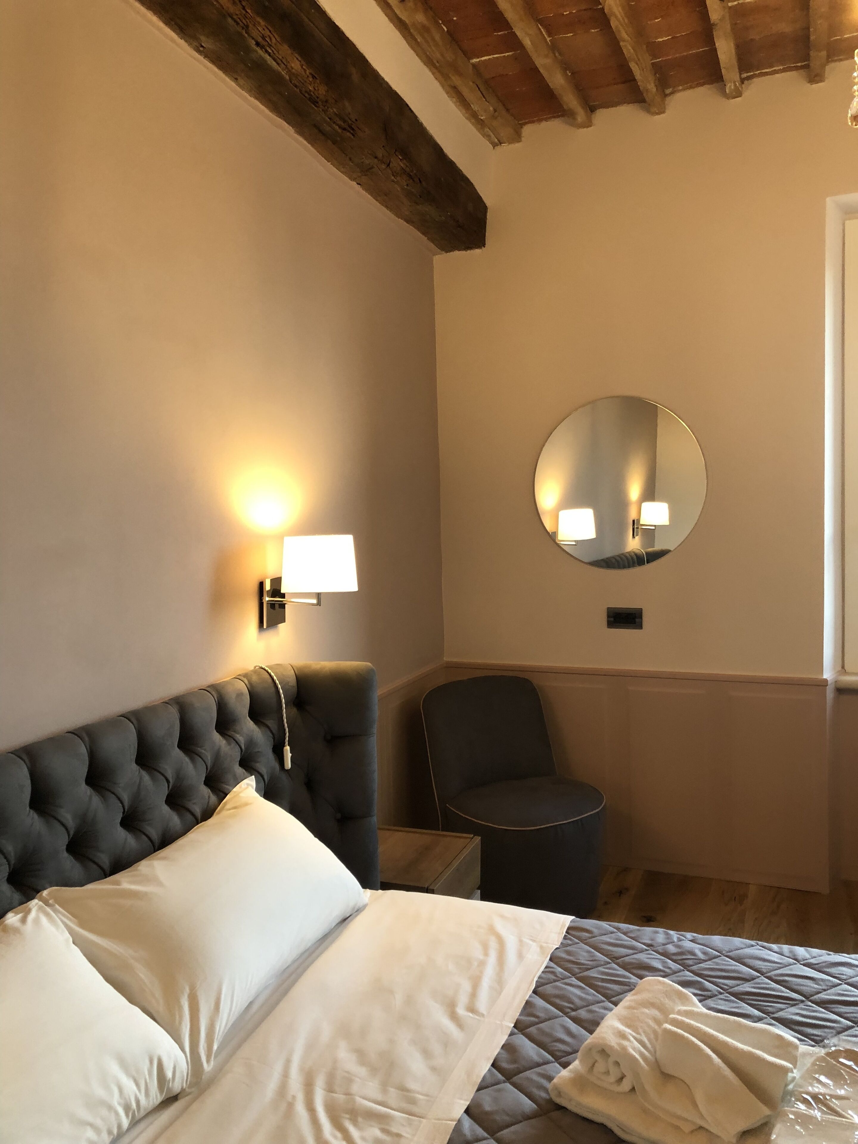 A hotel room with a tufted headboard, a round mirror, a bedside lamp, a chair, and a neatly made bed with a quilted cover.