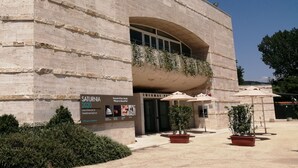 Un edificio moderno con fachada de piedra, un amplio acceso en arco y zona de estar exterior con sombrillas.