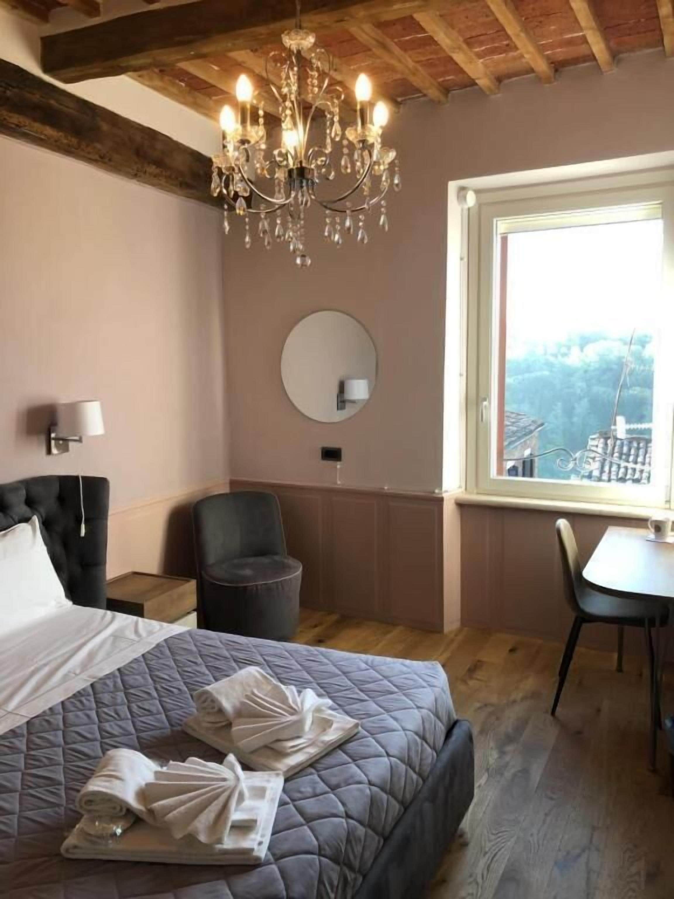 A bedroom with a large bed, a chandelier, a round mirror, a chair, and a table by a window with a view.