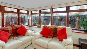 Una sala de estar acogedora con un sofá de cuero blanco, cojines rojos y vistas a un jardín a través de amplias ventanas.