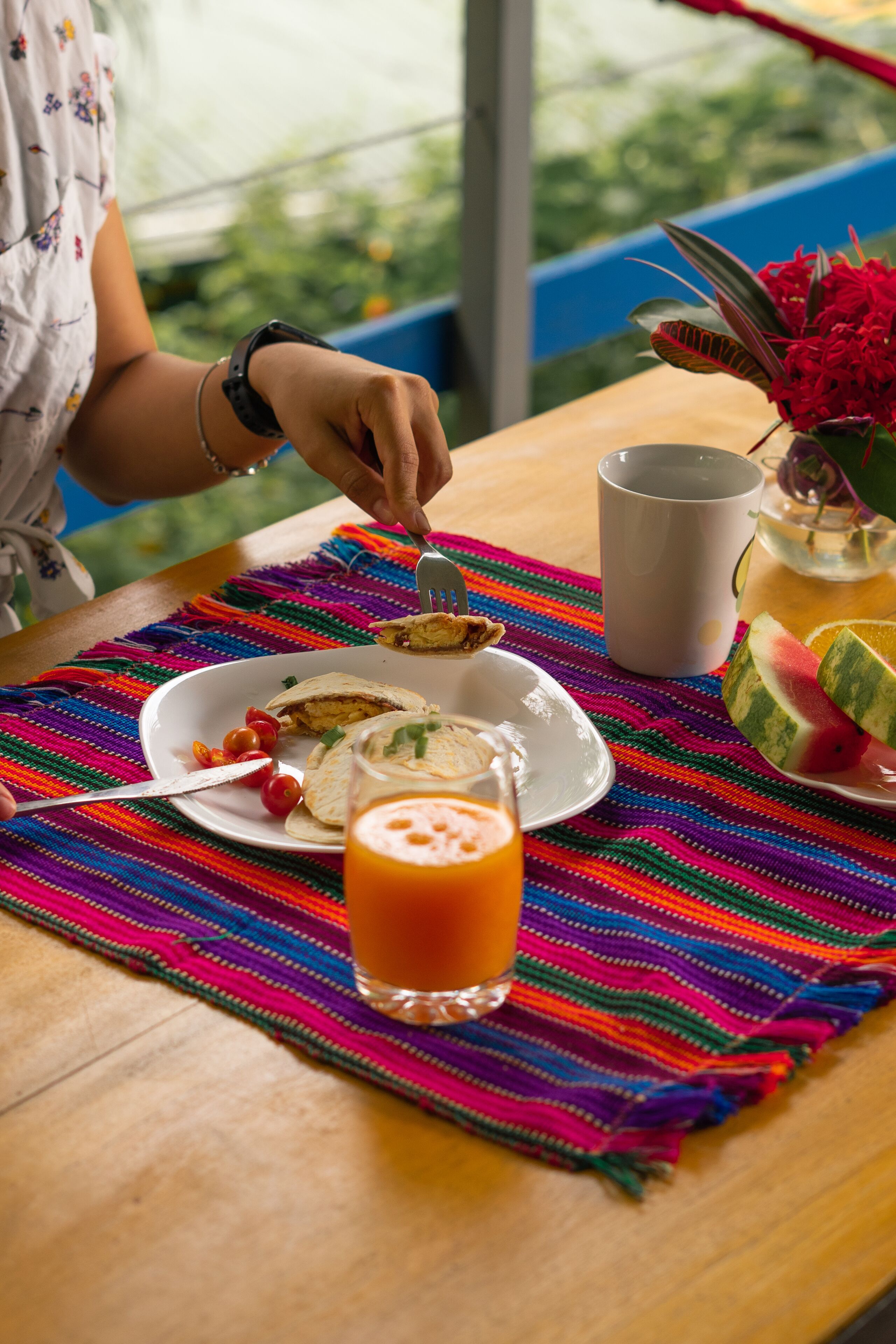 Una mesa con un posavasos tejido y de colores, un plato con comida, un vaso de jugo de naranja y una taza blanca.