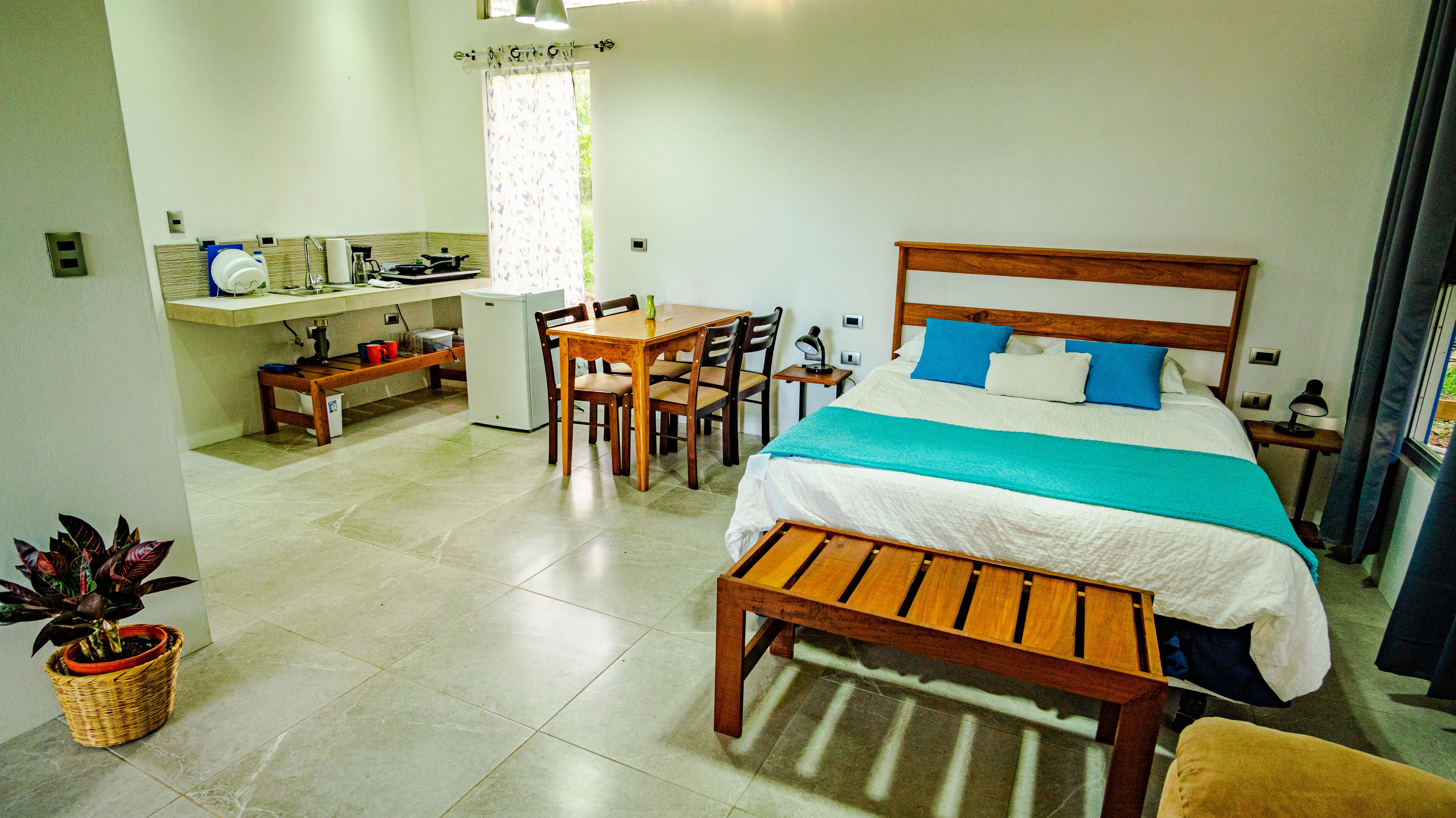 A bedroom with a bed, bedside table, lamp, and a view of the kitchen area.