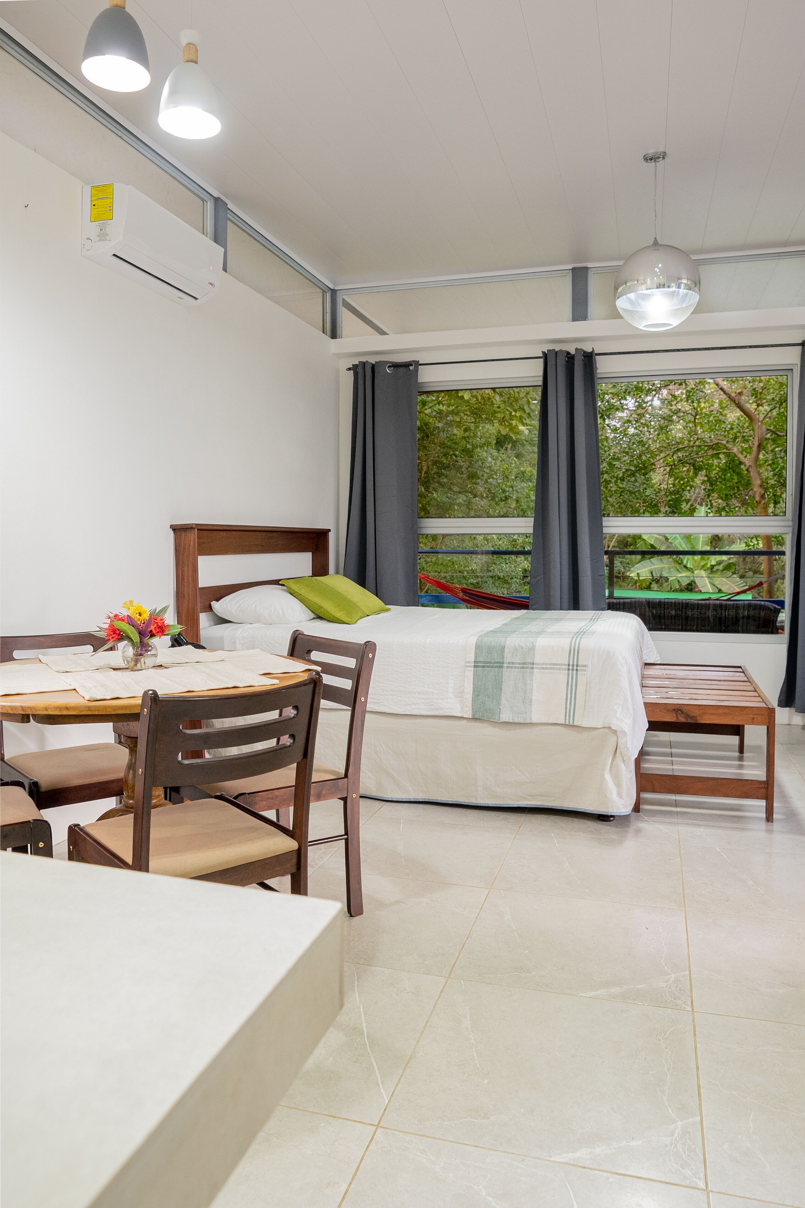 A hotel room with a bed, a dining table, chairs, a small table, and a view of greenery outside.