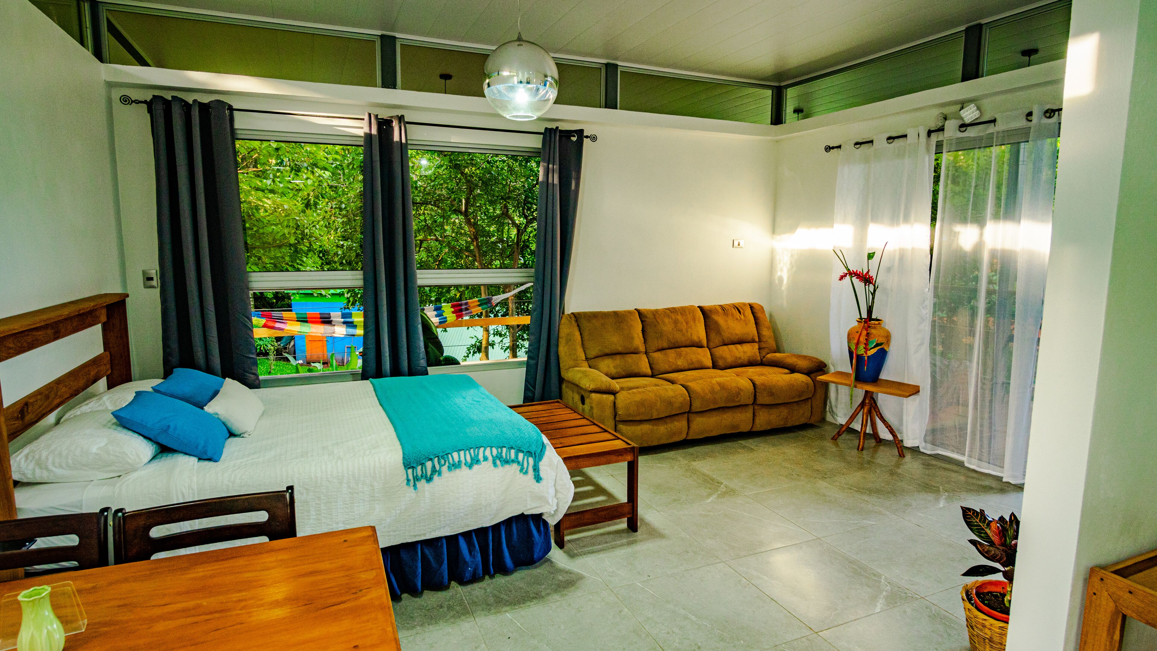 Quarto com cama, sofá, mesa pequena e janela com cortinas.