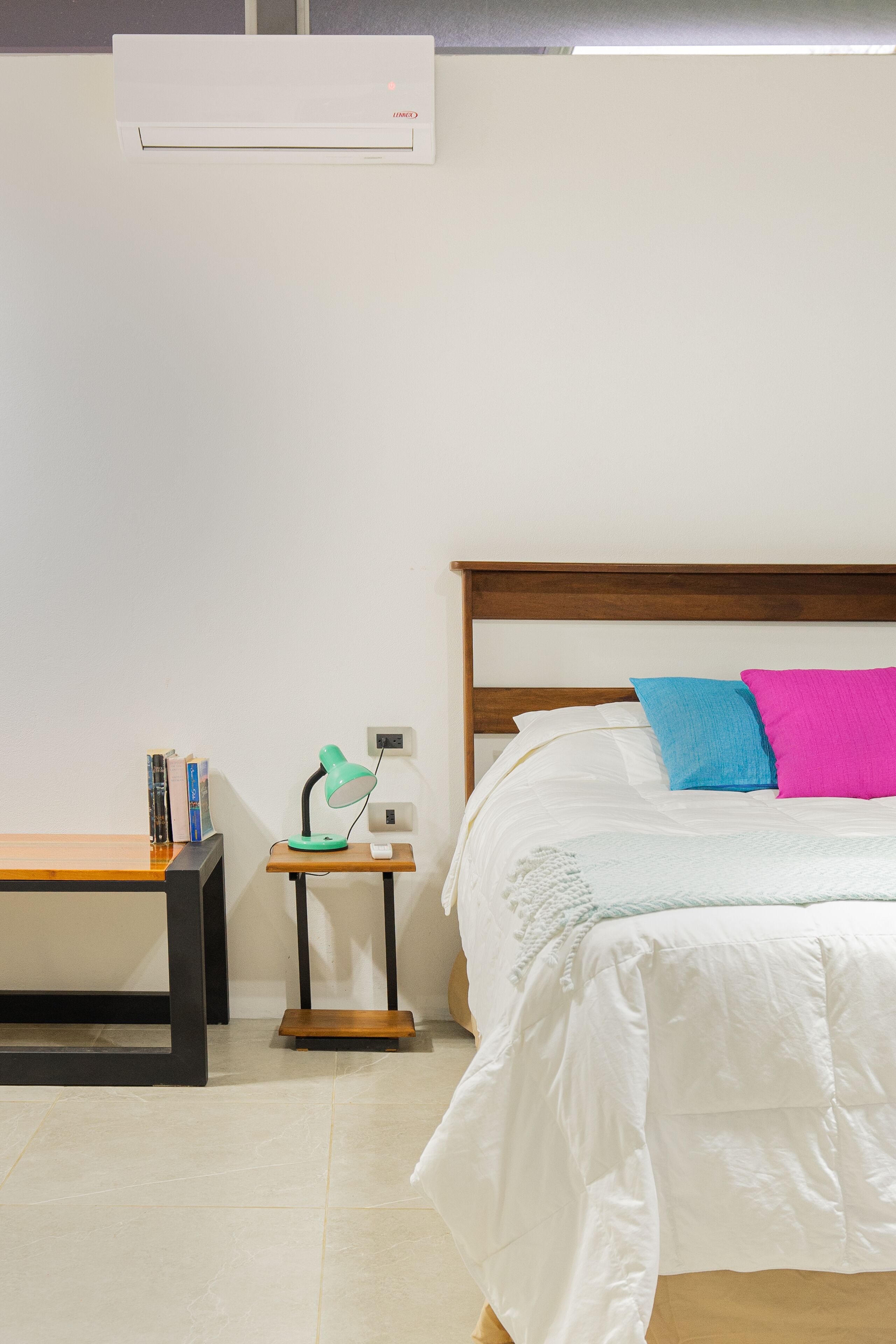 Een slaapkamer met een houten hoofdbord, een bed met witte beddengoed en twee kussens, een nachtkastje met een lamp, en een klein houten bankje.