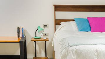 Een slaapkamer met een houten hoofdbord, een bed met witte beddengoed en twee kussens, een nachtkastje met een lamp, en een klein houten bankje.