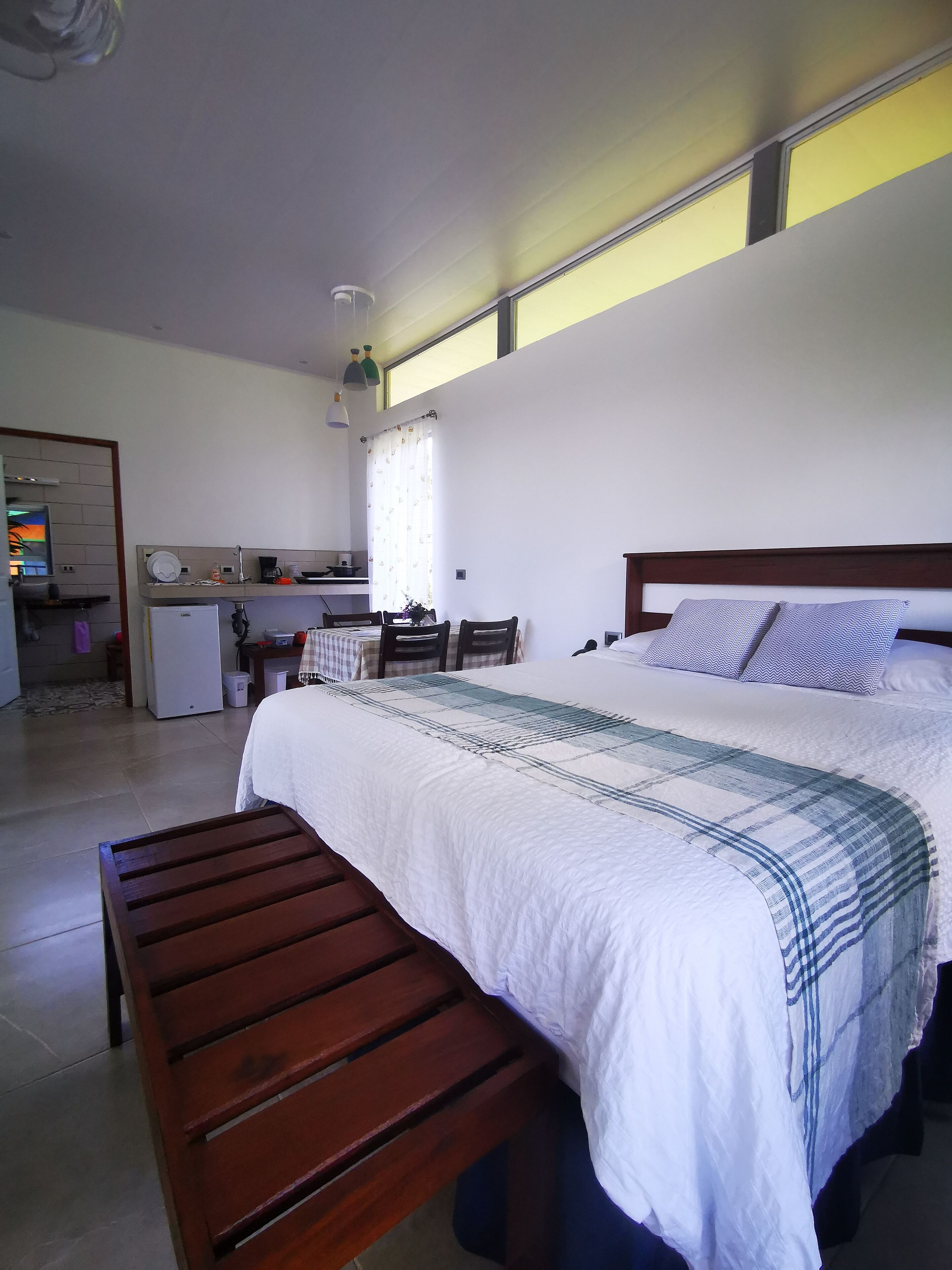 A bedroom with a large bed, a wooden bench, a small kitchenette, and a dining area.