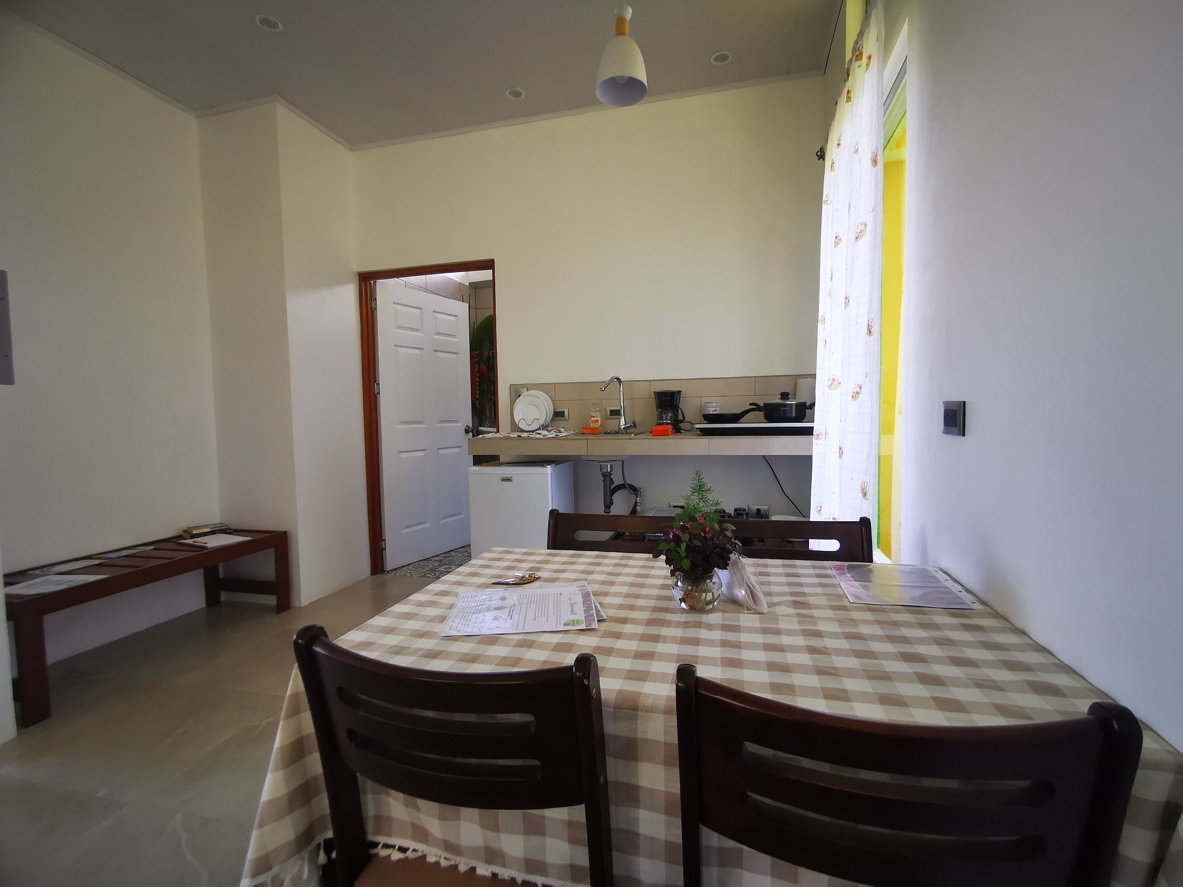 Un comedor con mantel a cuadros, sillas de madera y una pequeña planta en maceta. Se ve una cocina al fondo, con un refrigerador y un microondas.