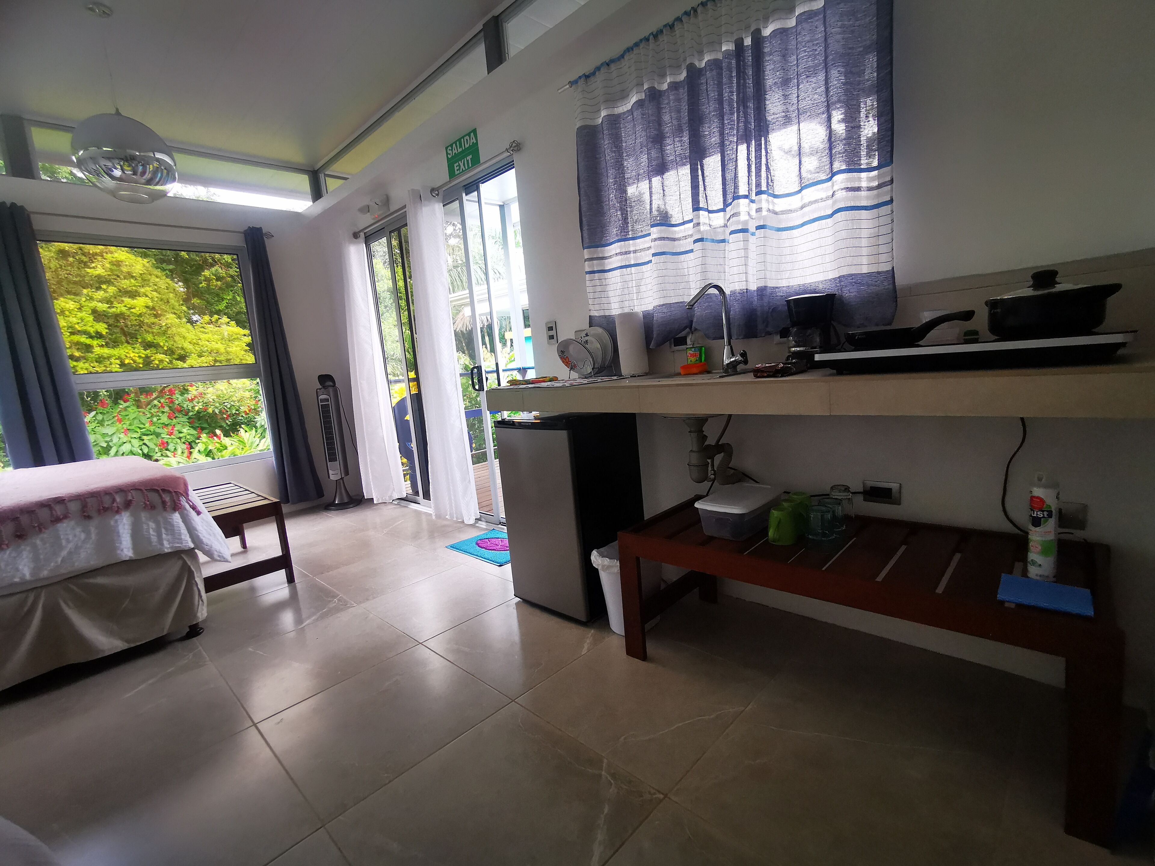 A room with a bed, a desk with a computer, a window with curtains, and a view of greenery outside.
