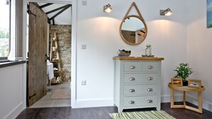A hallway with a wooden door, a grey dresser with drawers, a round mirror, and a small table with a plant and picture frame.