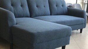 Ein blaues Ecksofa mit abgesteppten Rückenlehnen und dunklen Holzbeinen auf einem Fliesenboden.