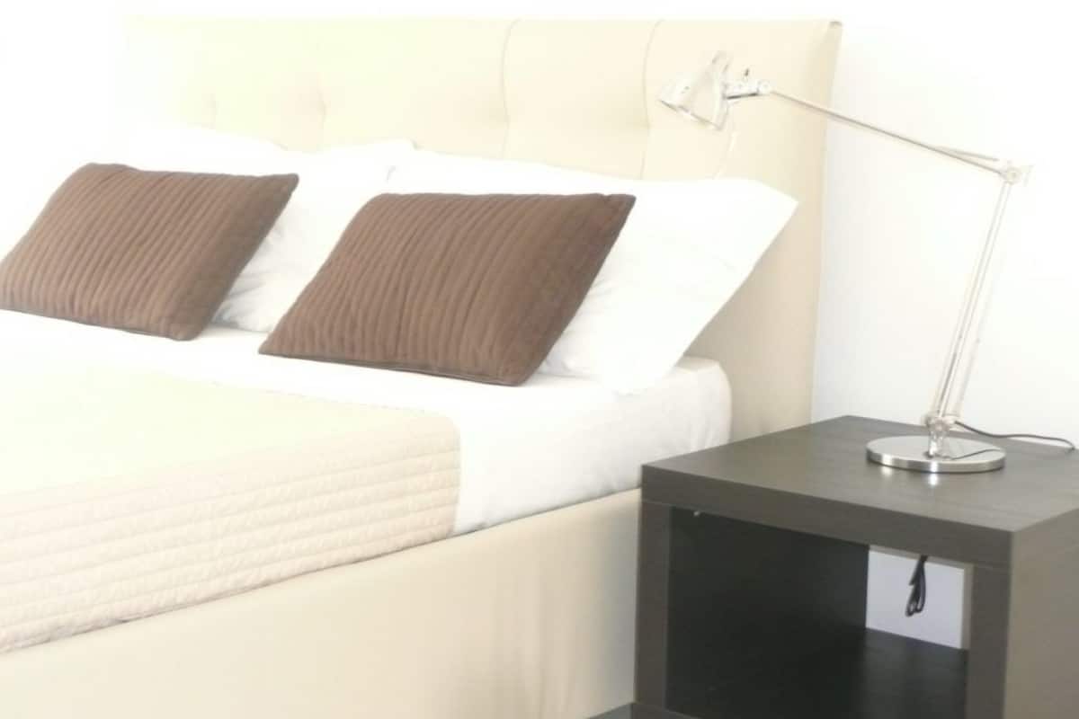 A bed with white linens and two brown pillows, a bedside table with a lamp, and a small drawer.