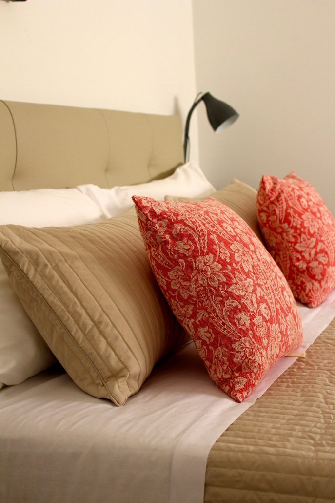 A bed with beige bedding and two red patterned pillows.