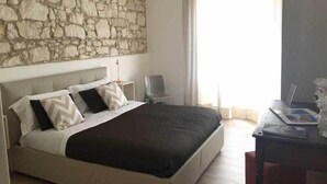 A bedroom with a stone wall, a bed with a dark headboard, a bedside table, a chair, and a desk with a lamp.