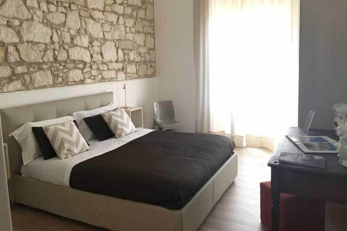 A bedroom with a stone wall, a bed with a dark headboard, a bedside table, a chair, and a desk with a lamp.