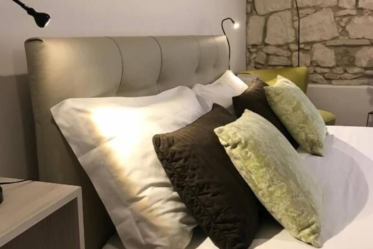 A bed with a headboard, multiple pillows, a nightstand, and a wall-mounted lamp.