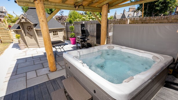 A hot tub in a backyard with a wooden deck, a barbecue grill, and a covered seating area. - Inverness (Inverness)
