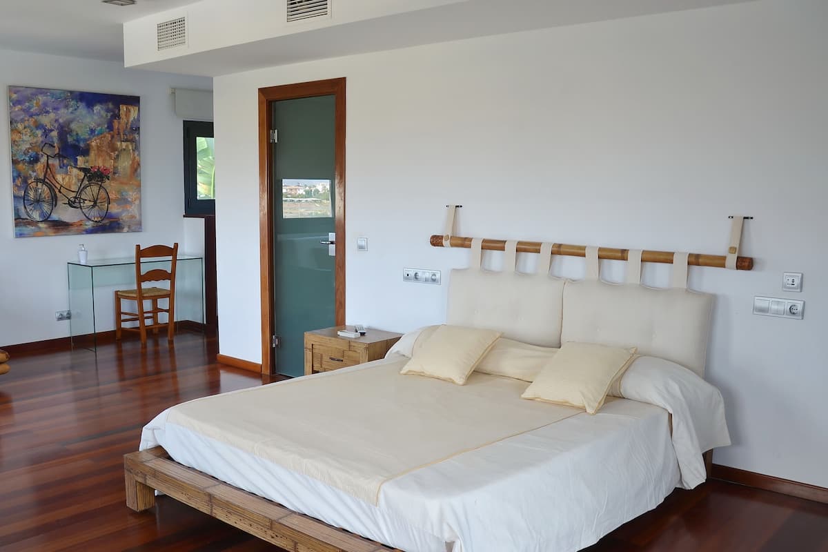 A bedroom with a wooden bed, a glass desk, a chair, and a painting on the wall.