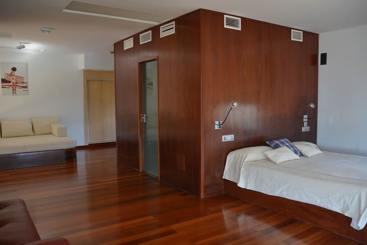 A bedroom with a wooden wardrobe, a bed with white bedding, a wooden floor, and a beige sofa.