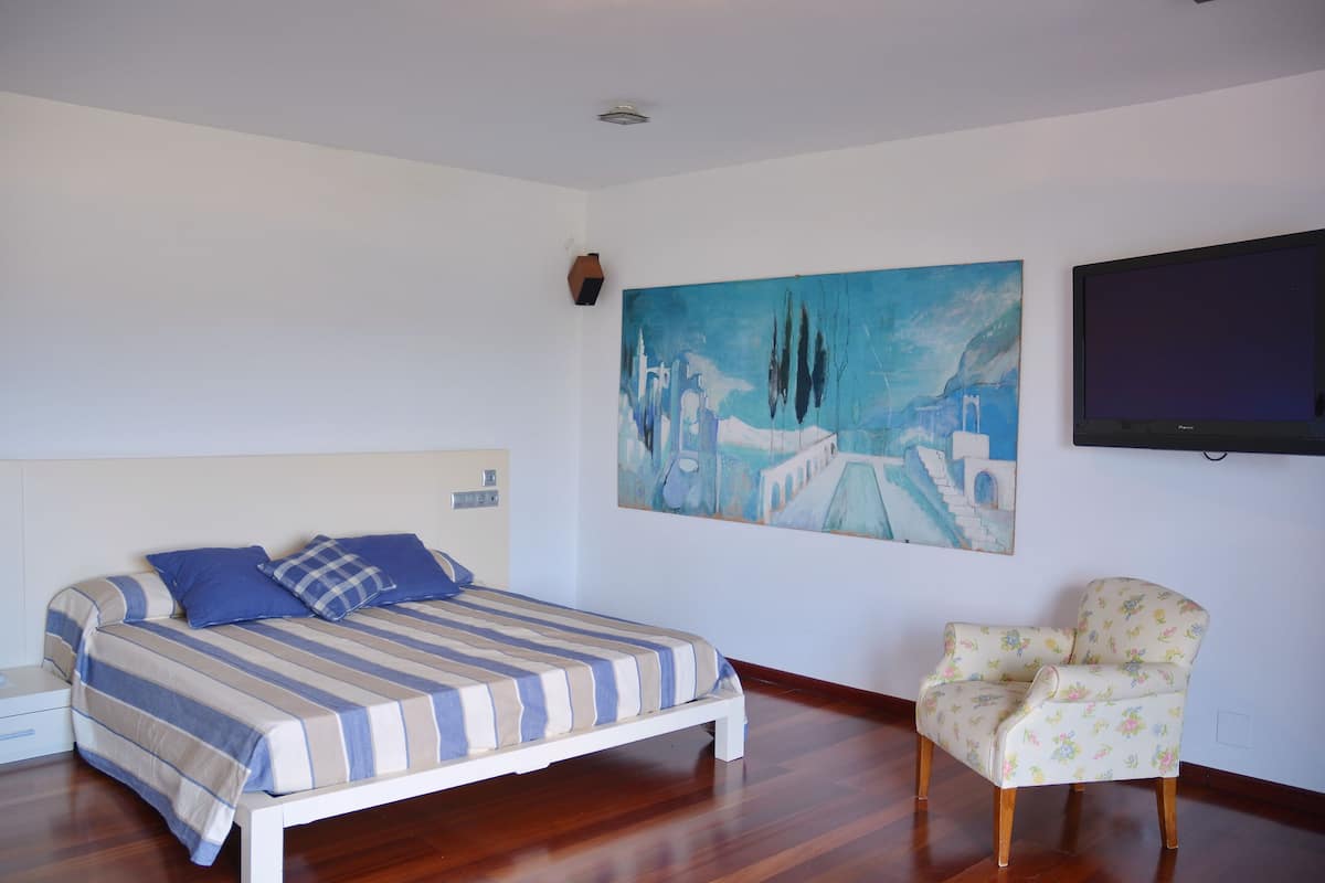 A bedroom with a bed, a chair, a painting, and a television.