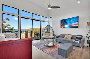 Ein geräumiges Wohnzimmer mit großen Fenstern, einem Deckenventilator, einem Stadtbildgemälde und einer modernen Sofagarnitur.