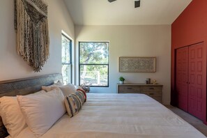 A bedroom with a large bed, a wooden headboard, a red door, a window with a view of trees, and a small plant on a nightstand. - Luxury Getaway - Waterfront, 4 BD, hot tub, stargazing. (Canyon Lake)