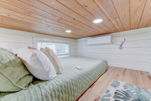 Quarto com cama, almofadas, janela com persianas, aparelho de ar condicionado e teto de madeira.