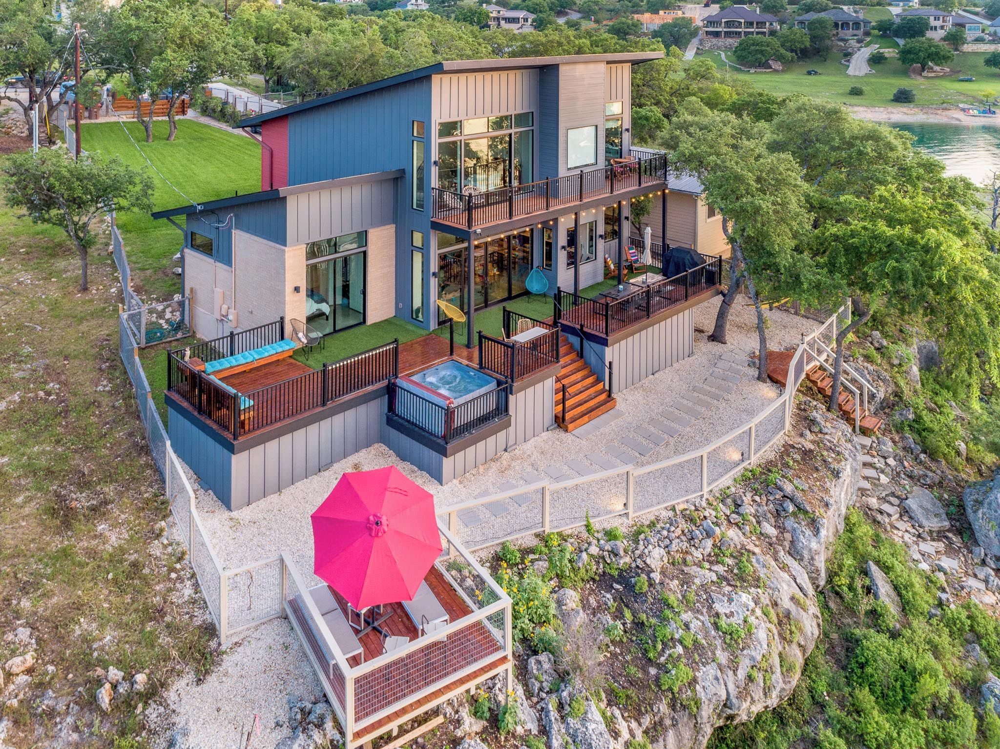 A modern two-story house with a hot tub, outdoor seating, and a deck overlooking a rocky area.
